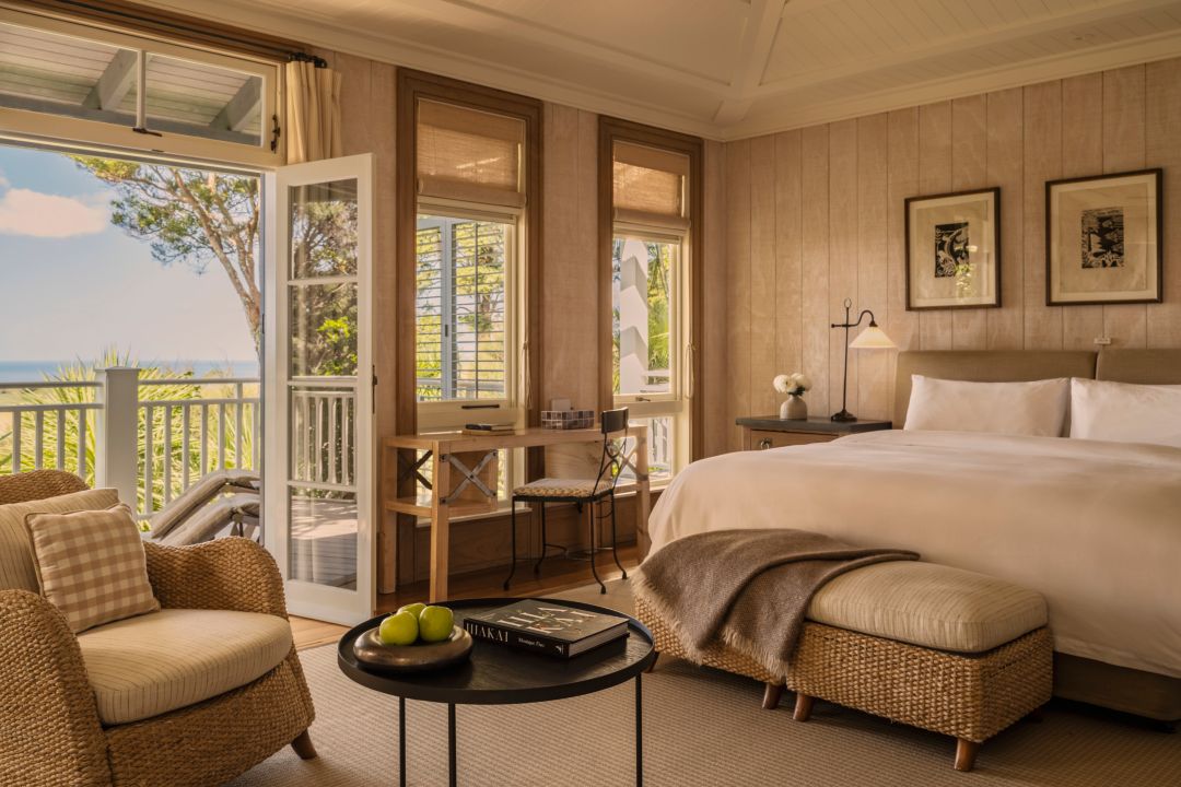 Spacious hotel suite with plush bed, armchair, desk and open doors out to a private balcony with golf course and ocean views.